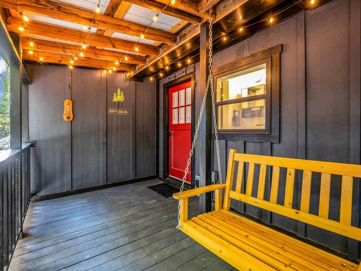 Finished Stonehaus covered porch with dark siding, red door, wooden swing and string lights